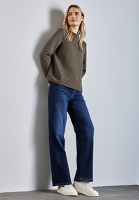 Oliivinvihreä tikkikuvioinen neule, jossa on pitkät hihat, yhdistettyinä väljiin tummansiniisiin farkkuihin ja valkoisiin tennareihin, neutraalilla seinää vasten.