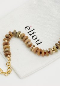 Bracciale di perline con pietre tonde multicolori in tonalità terrose, accentuato da parti in metallo dorato, su una pochette di tessuto bianco.