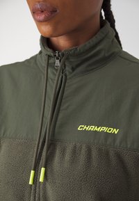 Giacca verde con zip, caratterizzata da una parte inferiore in morbido pile, una parte superiore liscia a contrasto e un prominente logo giallo "CHAMPION".