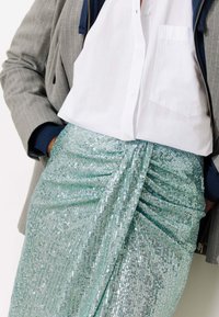 Jupe sequins bleu clair avec texture plissée, associée à une chemise blanche à boutons et un blazer gris à rayures.