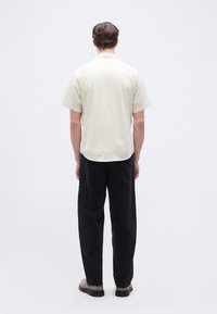 Άνδρας που στέκεται με την πλάτη προς τα εμπρός φοράει μπεζ ριγέ κοντομάνικο πουκάμισο, μαύρο παντελόνι και ανοιχτό γκρι παπούτσια σε λευκό φόντο.