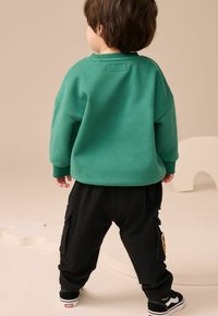 Sweatshirt vert surdimensionné avec des poignets côtelés, associé à un pantalon cargo noir ample avec des poches latérales et un patch animal. Baskets noires.