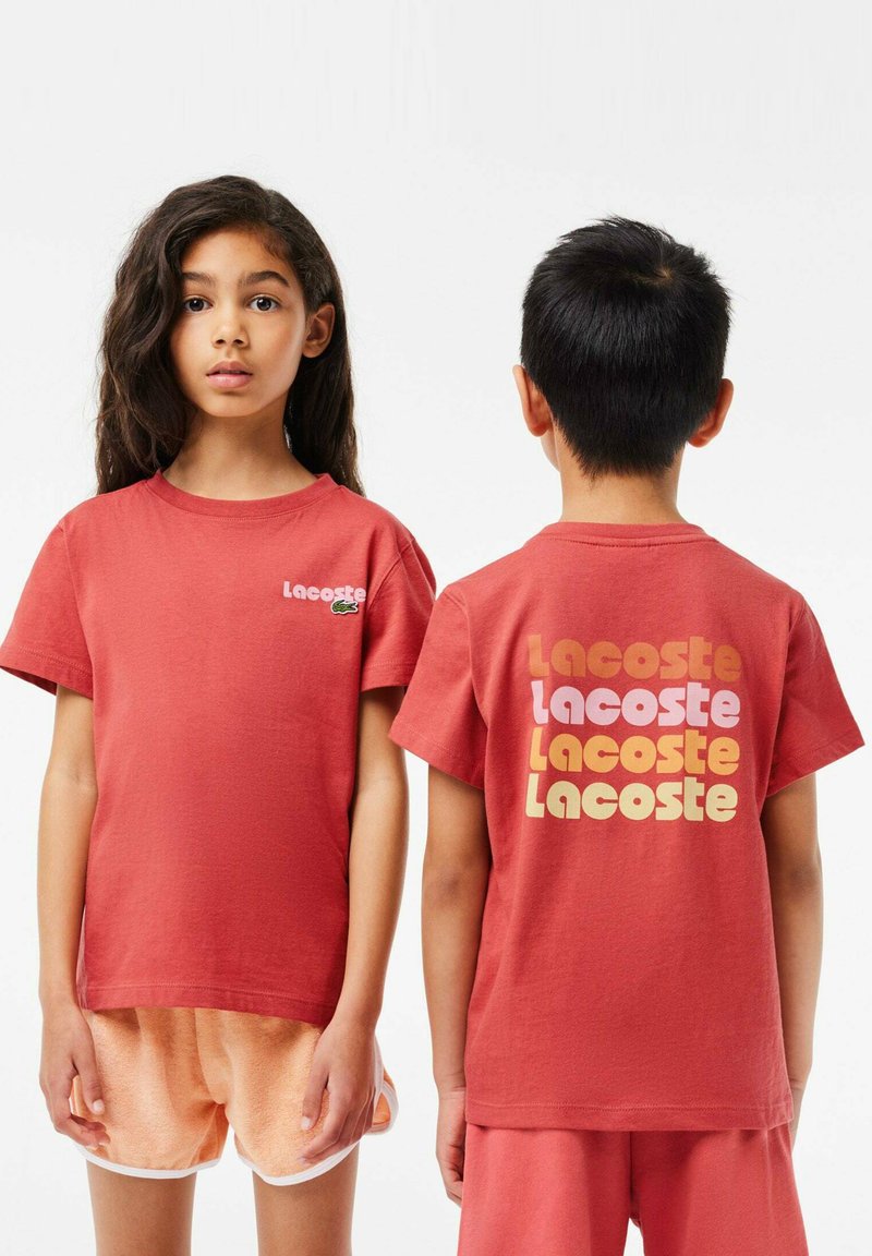 Rotes Baumwoll-T-Shirt mit kurzen Ärmeln. Mit Lacoste-Branding auf der Vorderseite und buntem Text auf der Rückseite. Lässiger Schnitt, glatte Textur.