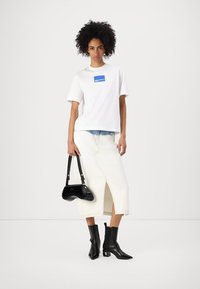 Tricou alb din bumbac cu un patch logo albastru, asortat cu o fustă albă din denim cu o fanta în față. Geantă de mână din patent negru și cizme cu gleznă.