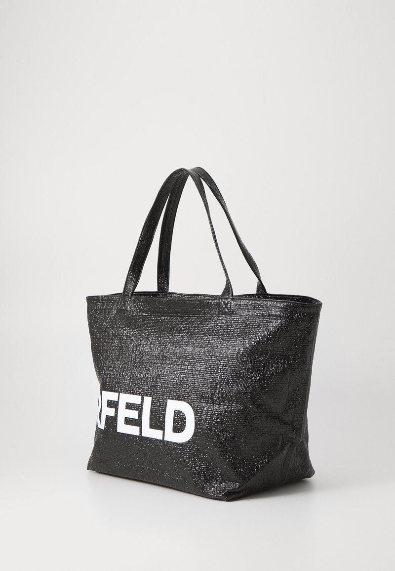 Black tote bag with a glossy texture, featuring two handles and a large white logo on the front. Rectangular shape, open top design.