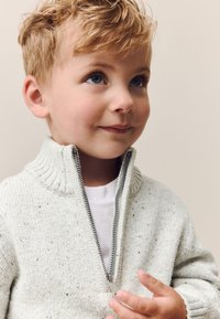 Strikket sweater, lys creme med små prikker, tekstureret stof, høj krave, lukning med halv lynlås, båret over en hvid skjorte, vist af et smilende barn.