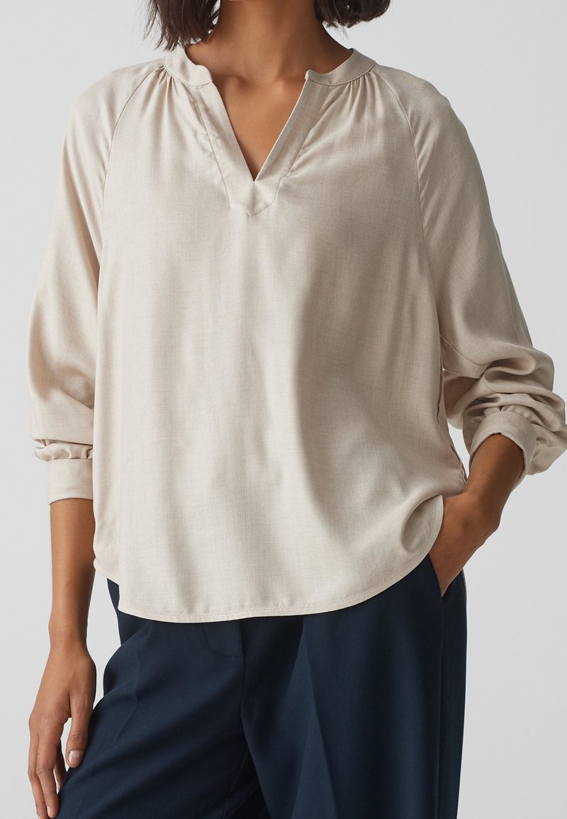 Femme portant un chemisier beige ample à manches longues avec un col en V et un pantalon bleu marine, une main dans la poche et l'autre détendue.
