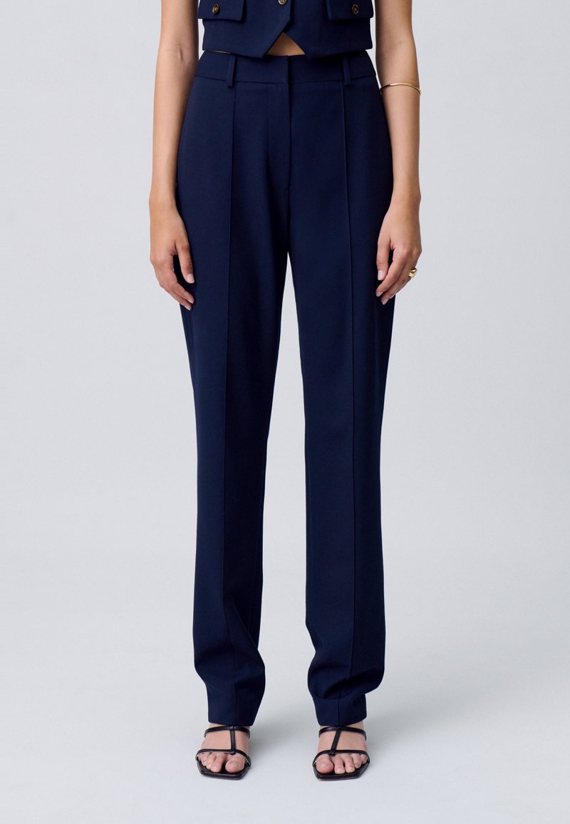 Modella che indossa un completo blu navy composto da pantaloni sartoriali e un gilet corto, in piedi con le mani ai fianchi su uno sfondo neutro.