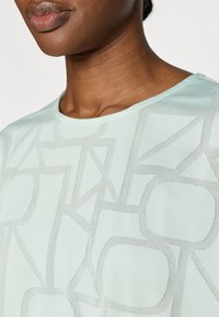 Ανοιχτό πράσινο τοπ με κοντά μανίκια και γεωμετρικό διατρητό μοτίβο, που περιλαμβάνει στρογγυλά και γωνι ώδη σχήματα, με λεία υφή και στρογγυλή λαιμόκοψη.