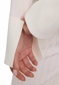 Witte gebreide top met geribbelde textuur, met een brede, licht opgerolde mouwzoom die een hand eronder onthult. Zacht, soepel materiaal.