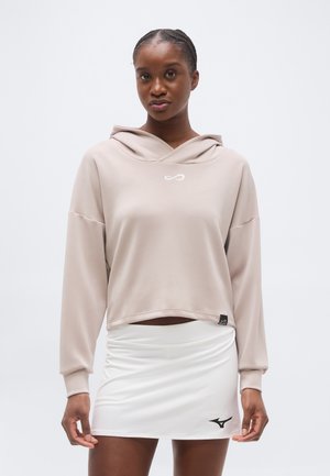 Femme portant un sweat à capuche court beige et une jupe de sport blanche avec un logo noir, debout devant un fond clair uni.