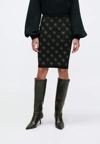 Schwarzer Bleistiftrock mit Diamantmuster und Logoakzenten, kombiniert mit kniehohen dunklen Lederstiefeln mit Absatz.