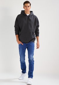 Kullsvart hettegenser med kenguru-lomme, parret med lyseblå jeans. Modellen har på seg hvite sneakers. Enkel, atletisk design.