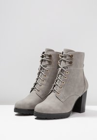 Paire de bottines en daim gris avec lacets sur le devant, œillets dorés et talon noir épais sur surface blanche contre un fond gris clair.