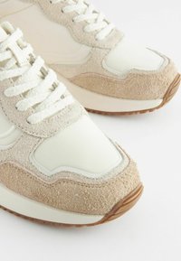 Las zapatillas de color claro presentan un upper liso y brillante con suaves acentos de ante, forma redondeada, superficie texturizada y suela de goma.