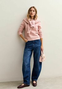 Lichtroze gebreide trui met een sjawlboord, gecombineerd met losse blauwe spijkerbroeken en bruine ballerina's, terwijl ze een kleine roze handtas vasthoudt.