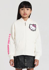Felpa zip con cappuccio di colore crema con grafica di Hello Kitty sul petto, caratterizzata da un fiocco rosa e il testo "FOLLOW ME"; tessuto morbido, vestibilità rilassata, due tasche.