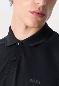 Μαύρο πόλο μπλουζάκι με γιακά, υφασμάτινη υφή, τρία κουμπιά και εκτύπωση του λογότυπου "BOSS" στο στήθος.