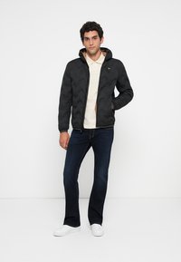Mand står iført en sort quilted jakke, beige polo skjorte, mørkeblå jeans og hvide sneakers mod en ensfarvet hvid baggrund.