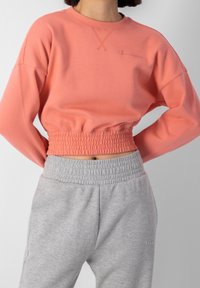 Kvinna i en korallfärgad elastisk kort sweatshirt och grå elastiska midje sweatpants, stående med händerna bakom ryggen.