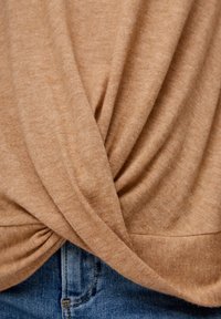 Beige Strickoberteil mit einem verdrehten Vorderdesign, glatter Textur und weichem Stoff, kombiniert mit blauen Denim-Jeans auf Taillenhöhe.