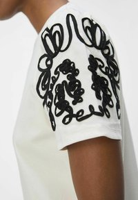 Vit bomull t-shirt med svarta broderade dekorativa detaljer på ärmarna. Tyget ser slätt och lätt ut.