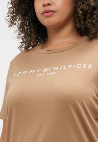 Rudas medvilninis marškinėlis su apvalia iškirpte ir trumpomis rankovėmis. Priekyje esantis grafikas rodo tekstą "TOMMY HILFIGER EST. 1985" baltomis raidėmis.
