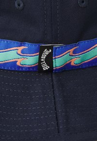 Námořnická čepice s modro-zeleným páskem ve vlnkovaném vzoru, s černou etiketou Billabong. Textura ukazuje jemné švy a odolný materiál.