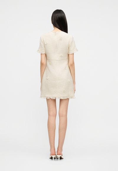 Γυναίκα με μακριά σκούρα μαλλιά φοράει κοντομάνικο, μπεζ φόρεμα με υφή, διακοσμημένο με λουλουδάτα σχέδια και φουντωτό τελείωμα, συνδυασμένο με μπεζ παπούτσια στιλέτο.