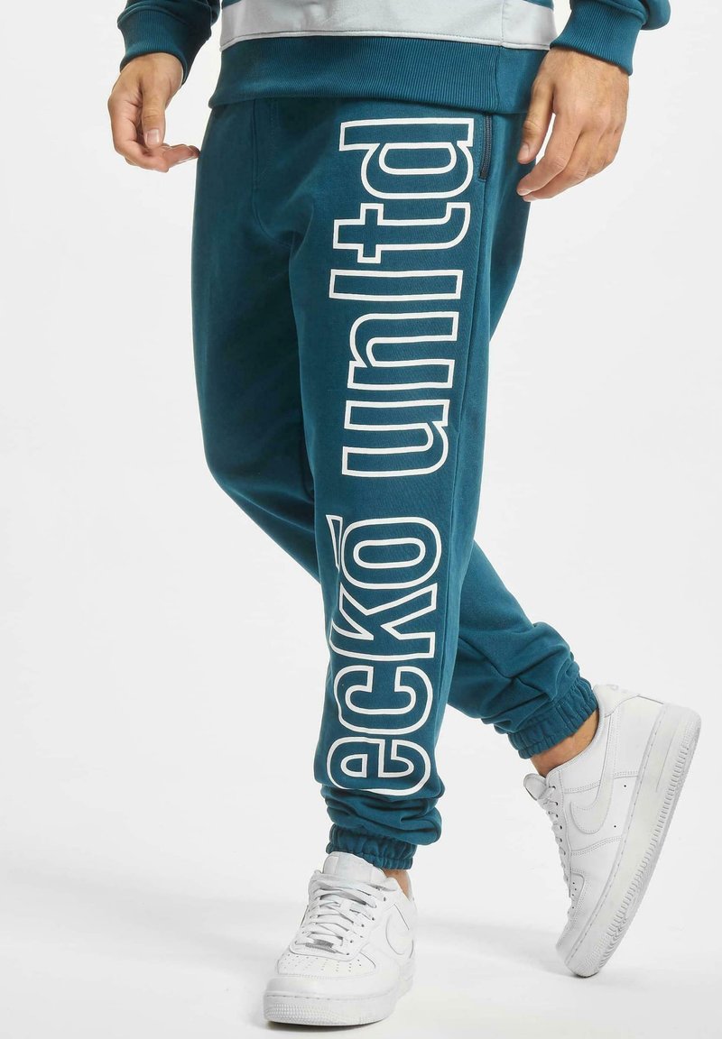 Teal joggebukser med en stor hvit logo trykket vertikalt på venstre bein, og elastiske manchetter ved anklene, parret med hvite sneakers.