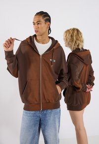 Brown zip-up hoodie with a gradient effect, front pockets, and a small logo. Fabric appears soft with a relaxed fit. Paired with light blue jeans.