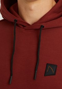 Acercamiento de una sudadera color burdeos con cordones negros y un pequeño parche de logo negro con un diseño naranja.