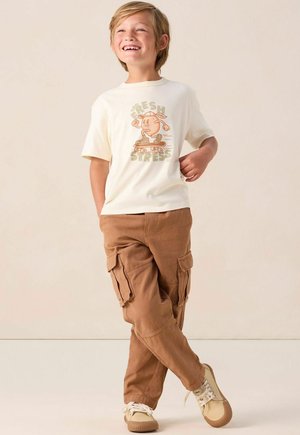 Cremefarbenes T-Shirt mit einem Grafikdesign eines Charakters auf einem Skateboard, kombiniert mit braunen Cargo-Hosen und beigen Sneakern mit weißen Schnürsenkeln.