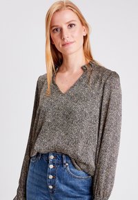 Blusa a maniche lunghe in tessuto scuro con motivo floreale bianco e oro. Presenta una scollatura a V e maniche arricciate. Abbinata a jeans blu a vita alta.