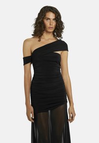 Vrouw met krullend haar in een zwarte, gerimpelde jurk met één schouder, voorzien van een doorschijnende stoffen overlay op de rok, staand tegen een effen achtergrond.