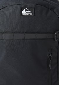 Schwarzer Rucksack mit Quiksilver-Logo, strapazierfähigem Stoff, strukturiertem Außenmaterial, vertikalen Trägern und einer Reißverschlusstasche an der Seite.