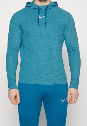 Mężczyzna ubrany w turkusową bluzę z kapturem Nike ze sznurkami i turkusowe sportowe spodnie Nike, stojący na tle jasnego, jednolitego tła.