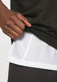 Maglietta sportiva nera con superficie testurizzata, sovrapposta a uno strato sottostante in rete bianca, che mette in evidenza tessuti contrastanti e un design complessivo traspirante.