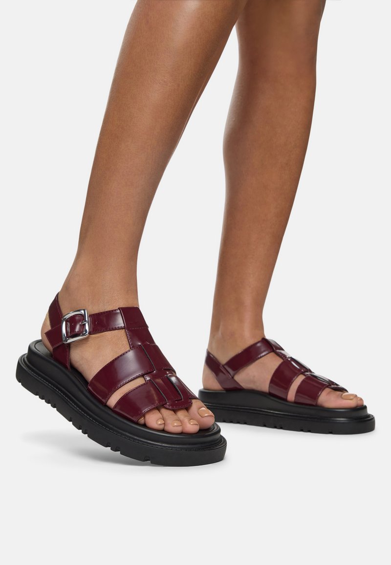 Burgundy patent leather sandals with thick black rubber soles. Features include straps with a buckle, an open-toe design, and grid pattern.