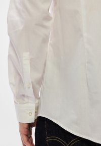 Λευκό βαμβακερό πουκάμισο με μανσέτα με κουμπιά, πτυχές στα πλάγια και λείο ύφασμα, σε συνδυασμό με σκούρο μπλε τζιν.