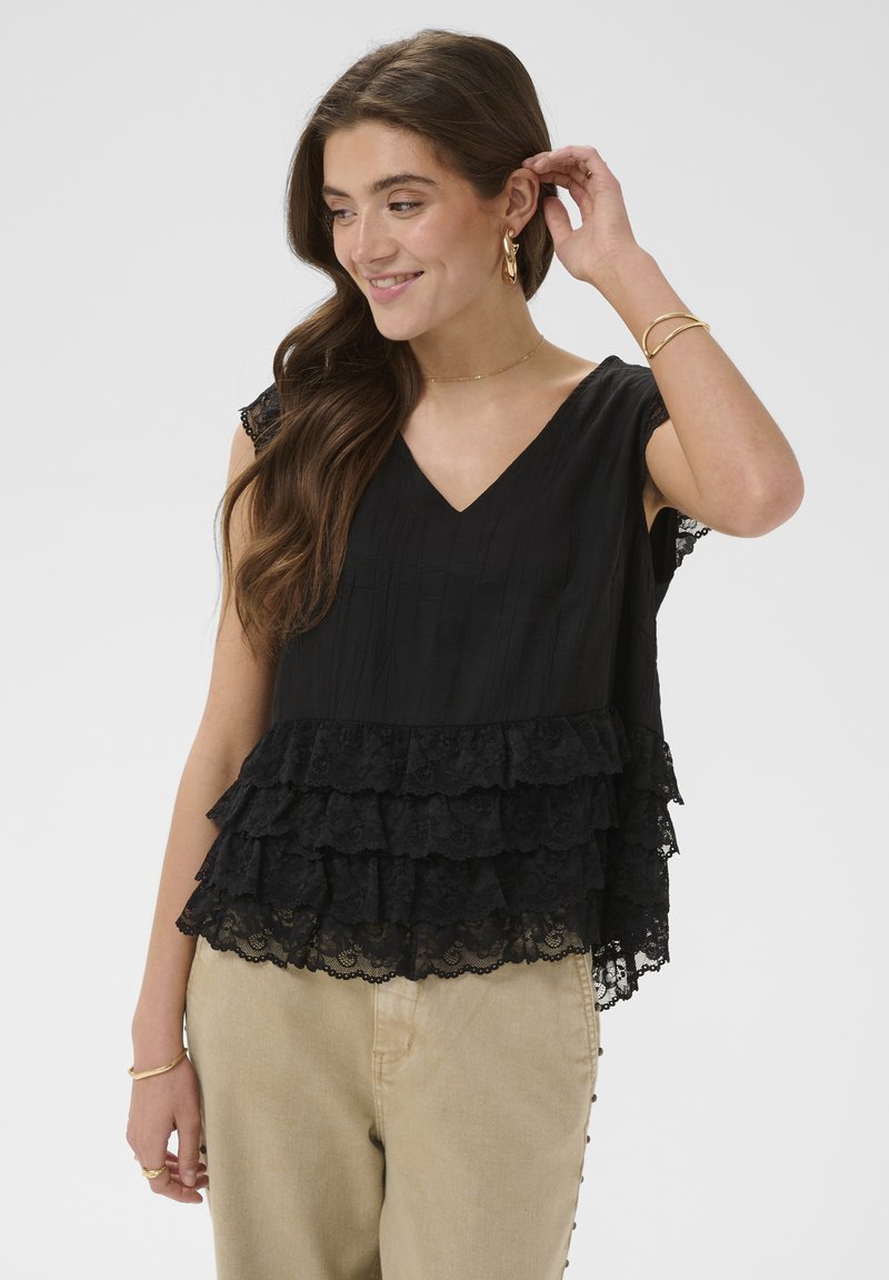Jeune femme aux longs cheveux bruns portant des boucles d'oreilles créoles en or, un haut en dentelle noire à volants superposés et un pantalon beige, souriante et touchant ses cheveux.