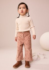 Pantaloni in corda color sabbia con motivi a fiocco, abbinati a un maglione a collo alto a coste color crema. Stivali alla caviglia marroni completano l'outfit.