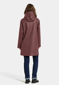 Waterdichte kastanjebruine regenjas met een grote capuchon, lange mouwen en een rechte snit. Draagtt over donkere jeans en zwarte enkellaarzen.