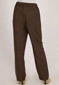 Lichte, losse bruine broek met een elastische tailleband en verticale naden, rustend boven open-hak schoenen met een gestructureerd oppervlak.