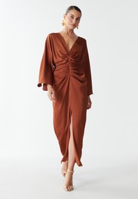 Rustfarvet maxi-kjole med dyb V-hals, rynket detalje ved taljen og lange ærmer. Har en frontslids for ekstra stil.