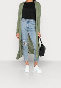 Largo cárdigan verde oliva sobre una camiseta negra, acompañado de jeans desgastados de color azul claro y zapatillas blancas. Incluye un bolso negro.
