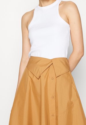 Kobieta ubrana w białą, bezrękawnikową, prążkowaną bluzkę wsuniętą w wysoką, beżową spódnicę trapezową z szerokim, spiczastym paskiem i guzikami z przodu.