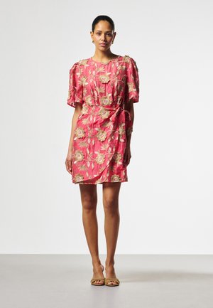 Frau steht in einem korallfarbenen geblümten Kleid mit Puffärmeln und Taillenschleife, trägt beigefarbene offene Absätze, vor einem einfarbigen Hintergrund.