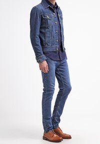 Giacca di jeans con bottoni argentati, due tasche sul petto, camicia blu con bottoni, jeans skinny blu e scarpe marroni stringate.