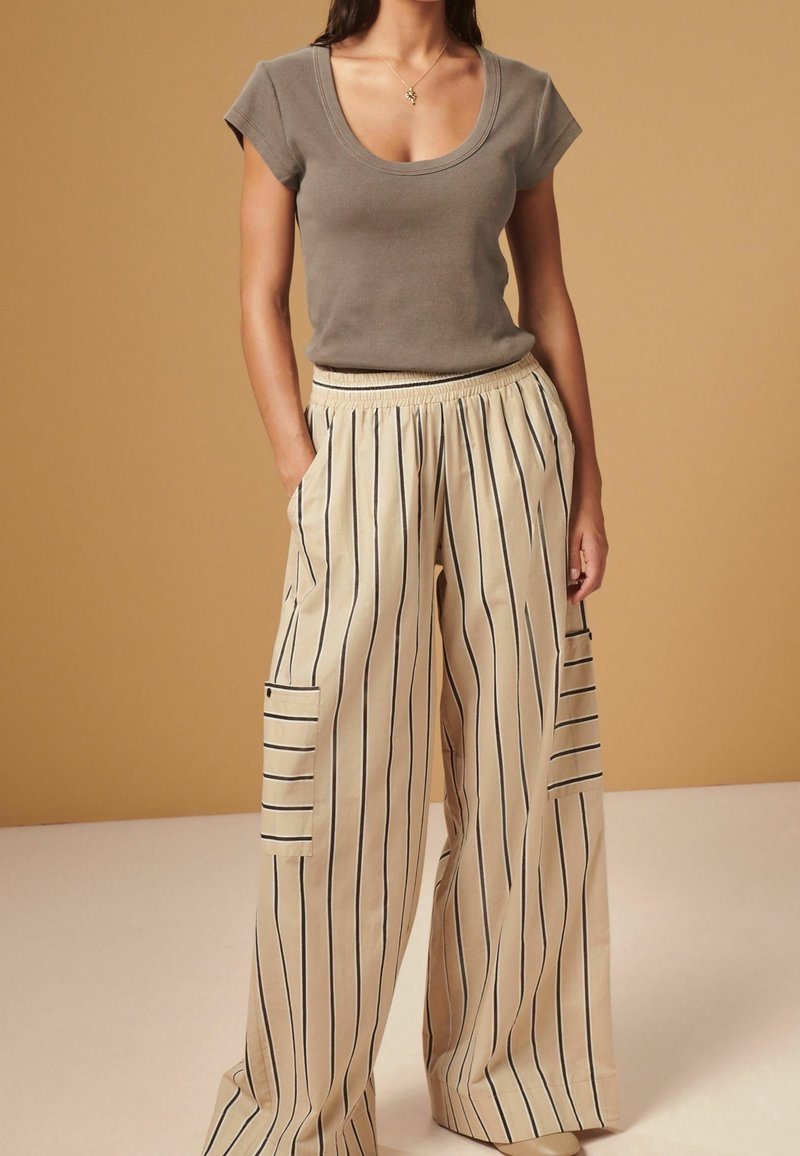 Femme portant un haut gris à col rond et manches courtes, ainsi qu'un pantalon beige à jambes larges avec des rayures noires verticales et des poches latérales, debout les mains dans les poches.
